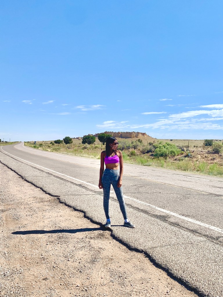 Jasmine standing in the middle of a dusty desert road. She wears a mask and sun glasses and stares off camera.