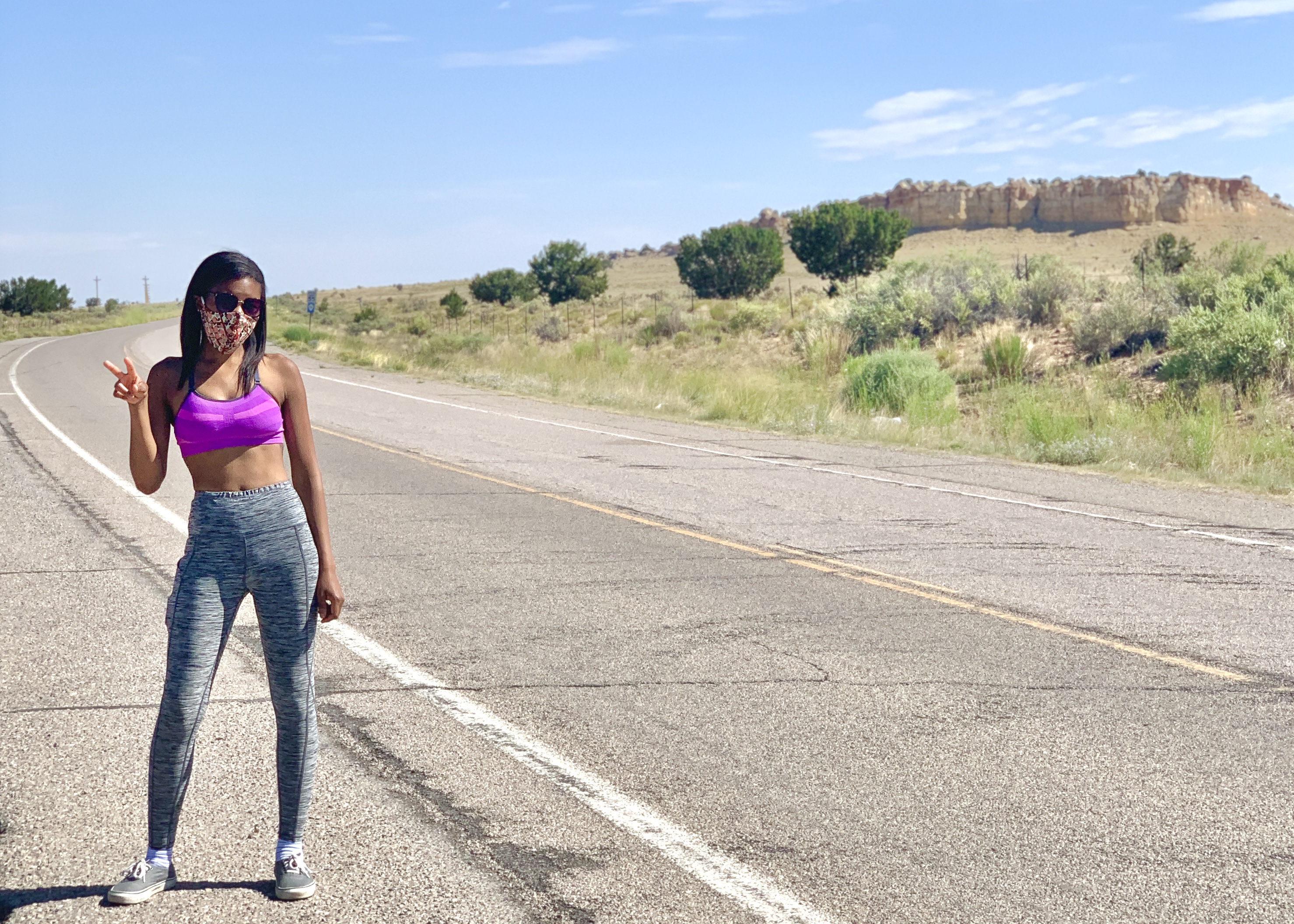 Jasmine wears a mask sunglasses and workout gear. She stands in the middle of an empty road flashing a peace sign.