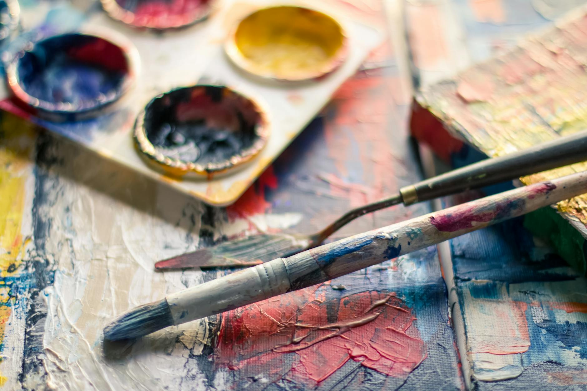 Image of paint, small pallet of used paints, a paint brush, and mixing tool on a wooden table covered in different colored paints.